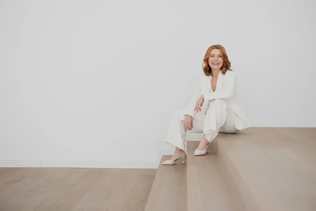 Karin Hübner im weißen Anzug sitzt lächelnd auf einer Holztreppe, Jobcoaching für Medienprofis