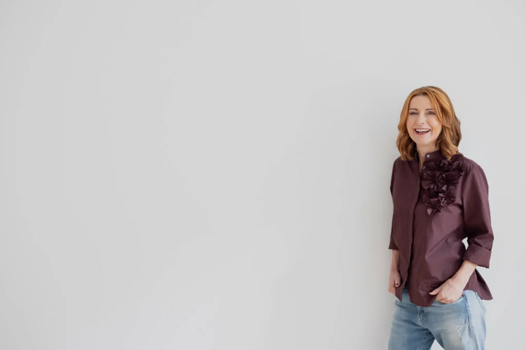Karin Hübner, Karriere-Coach aus Potsdam, steht lachend in Bluse und Jeans vor heller Wand